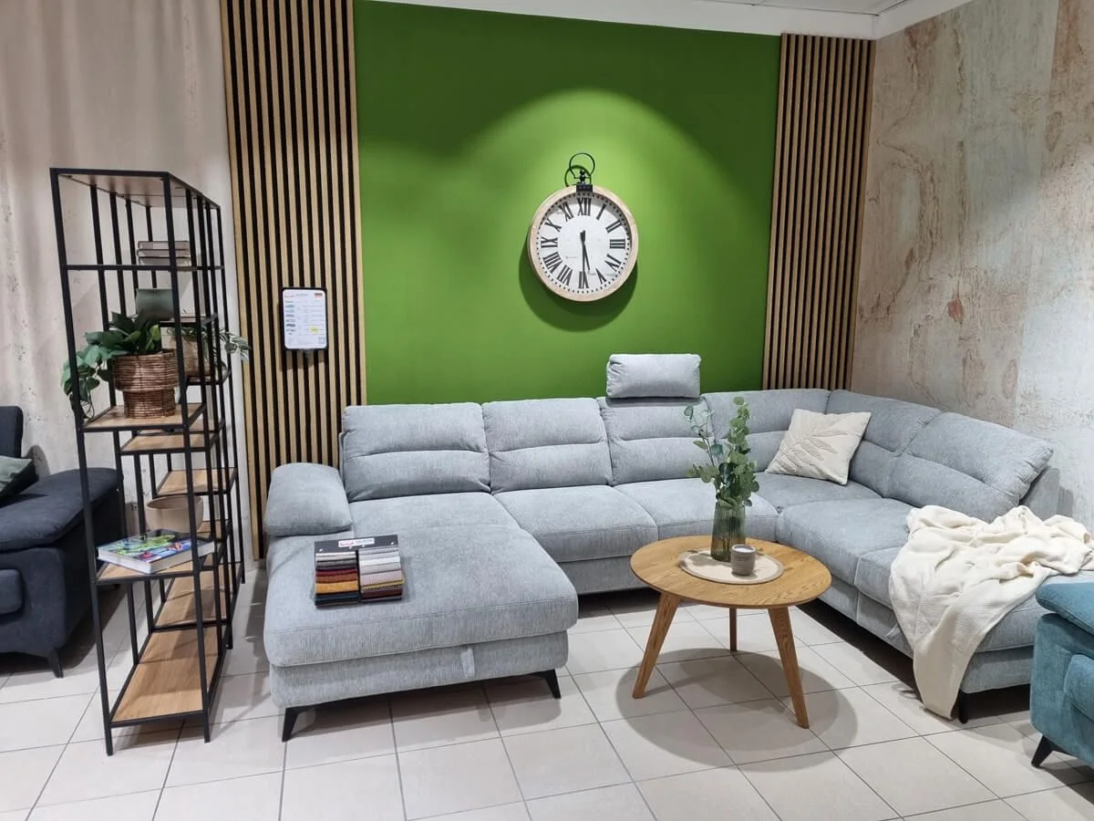 Šedá rohová sedací souprava v showroomu před zelenou stěnou s hodinami