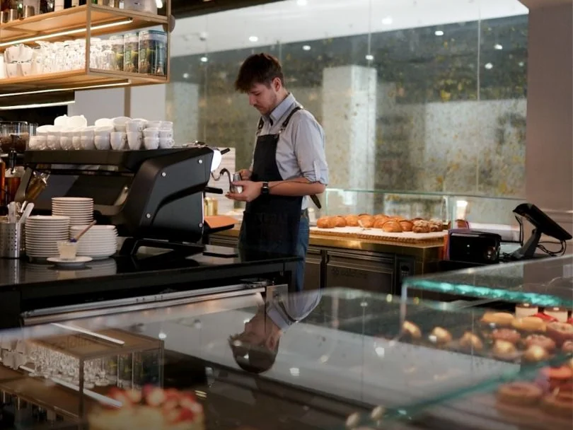 Barista u kávovaru za vitrínou s dezerty